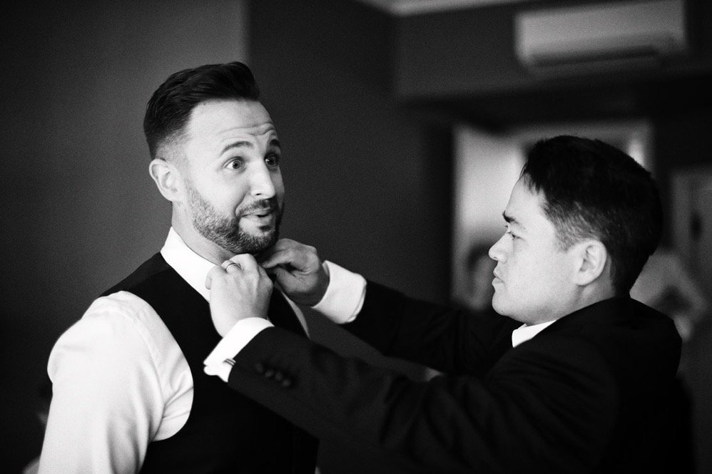 Two men are adjusting a bow tie on one man's neck in a black and white photo.