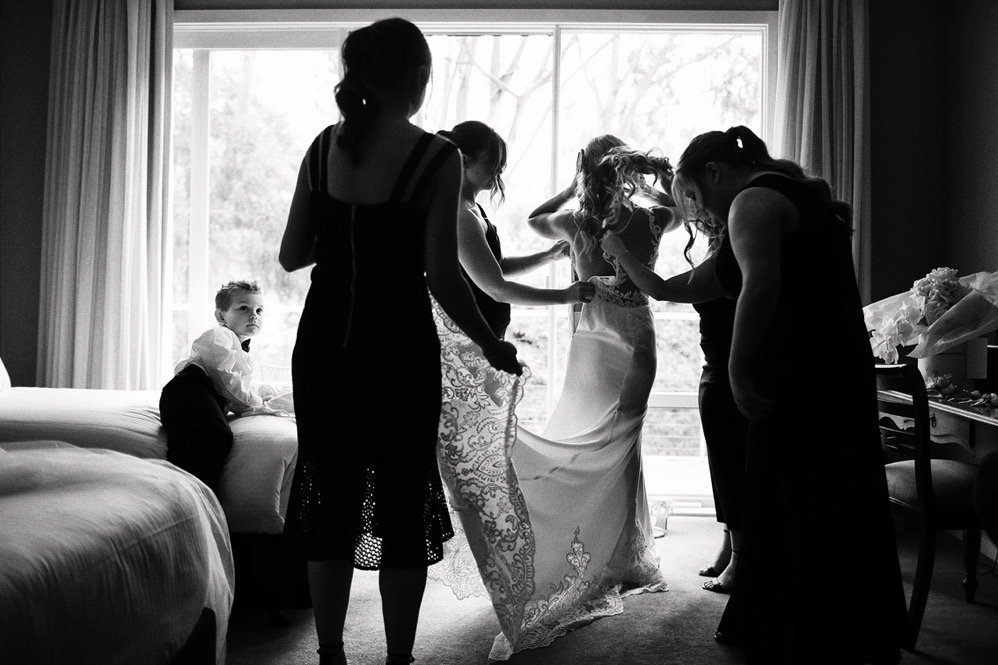 Women helping bride with her wedding dress in a room with large window, while children are sitting on beds.