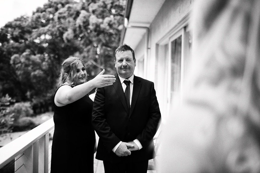 A woman is playfully pretending to shoot a man in a suit on a balcony with trees in the background.