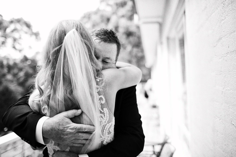 A bride and groom hug tightly during their wedding, with the bride wearing a veil and lace dress, and the groom in a black suit, outside on a porch.
