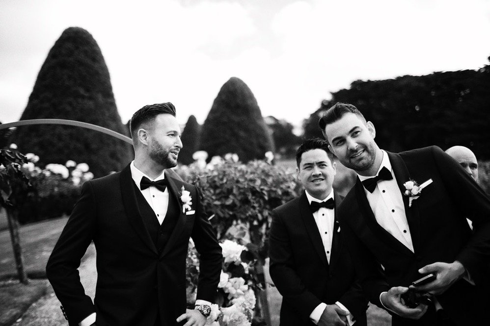 Three men dressed in tuxedos and bow ties at a wedding outdoors, with large, cone-shaped trees in the background, black and white photograph.