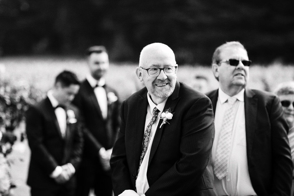 Smiling man in a suit at a formal outdoor event, with other men in suits and sunglasses in the background.