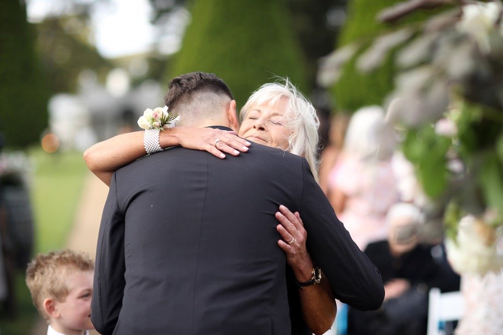 An elderly woman hugging a man in a suit, with a young boy nearby, in an outdoor setting during what appears to be a wedding or celebration, surrounded by blurred greenery and other guests.