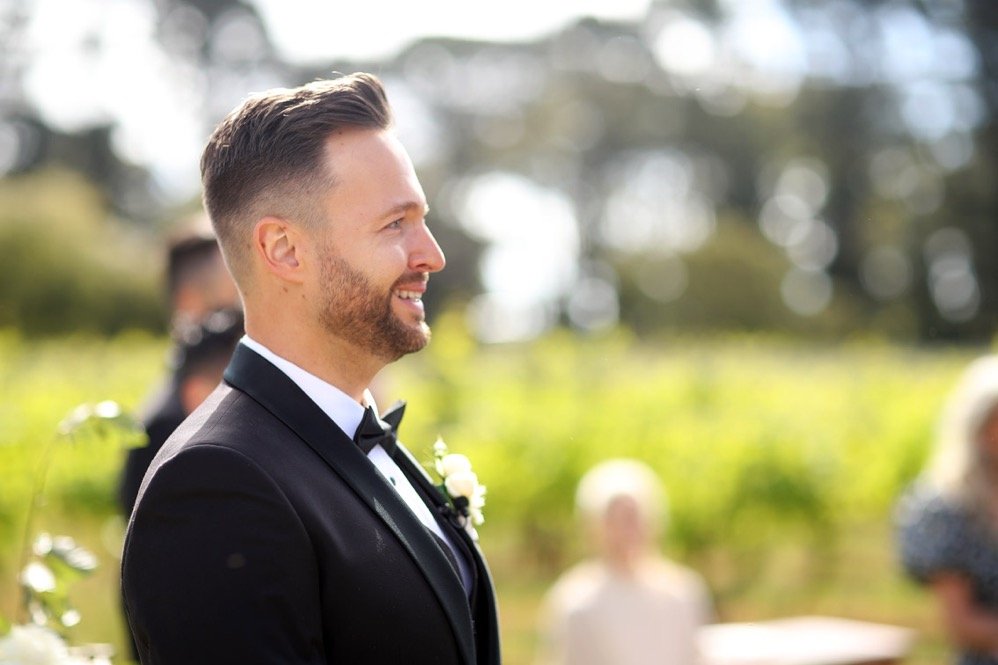 A man dressed in a black tuxedo with a white shirt and black bow tie, smiling at an outdoor wedding ceremony.
