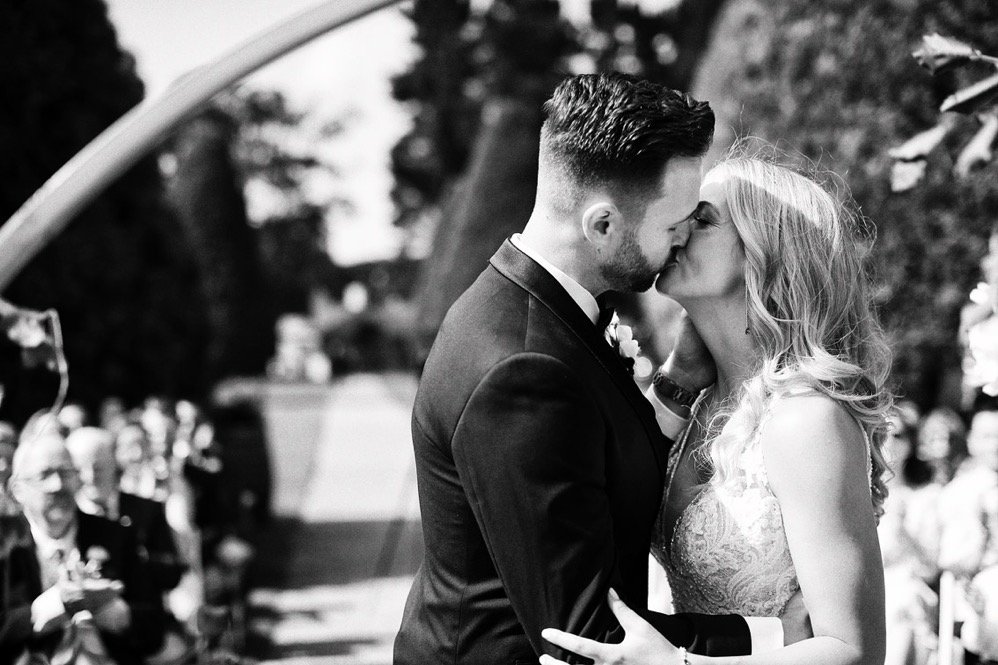 A newlywed couple sharing a kiss outdoors with guests watching in the background.
