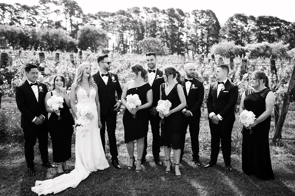 Black and white photograph of a wedding party outdoors, featuring a bride in a white gown, groom, and eight bridesmaids and groomsmen, all dressed in formal attire, standing on grass with trees and a vine-covered fence in the background.