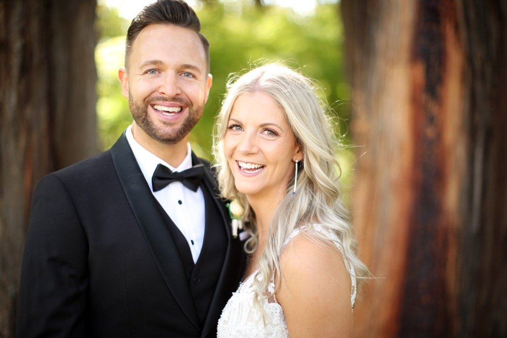 A smiling couple in wedding attire standing outdoors in front of trees, with sunlight filtering through the leaves.