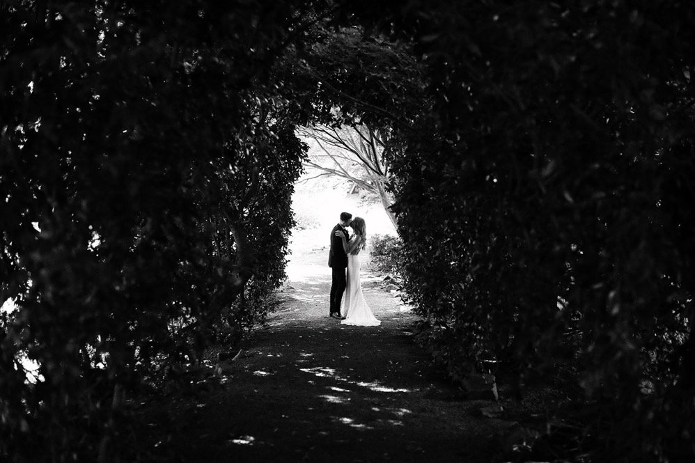 A couple in wedding attire sharing a kiss under a natural archway of bushes in a park setting.