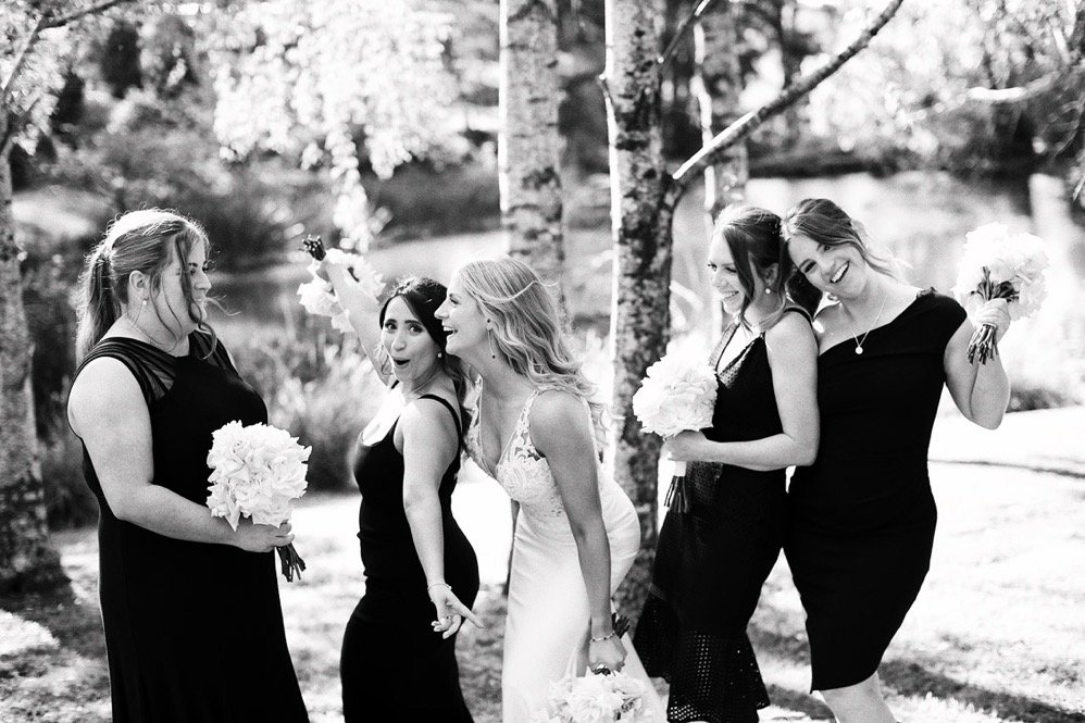 Group of five women, one in a wedding dress and four in black dresses, standing outdoors on grass with trees in the background, holding bouquets of flowers, celebrating and smiling.