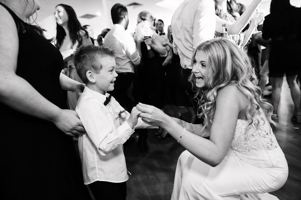 A woman in a wedding dress interacts happily with a young boy dressed in a shirt and bowtie at a festive event, possibly a wedding reception, with other guests dancing and socializing in the background.