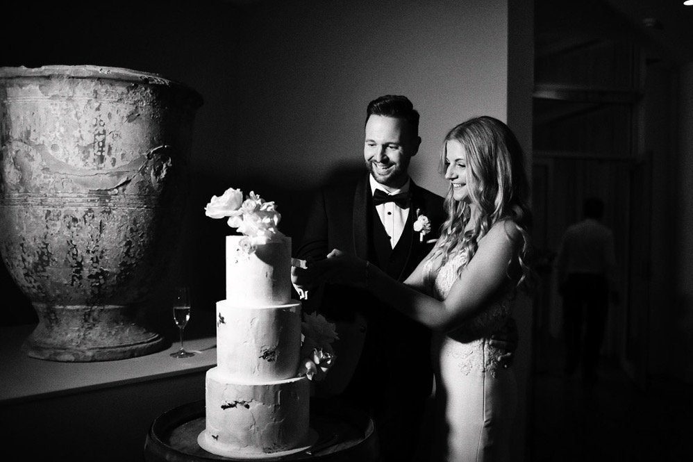 Black and white photo of a bride and groom cutting their wedding cake with a large decorative vase in the background.