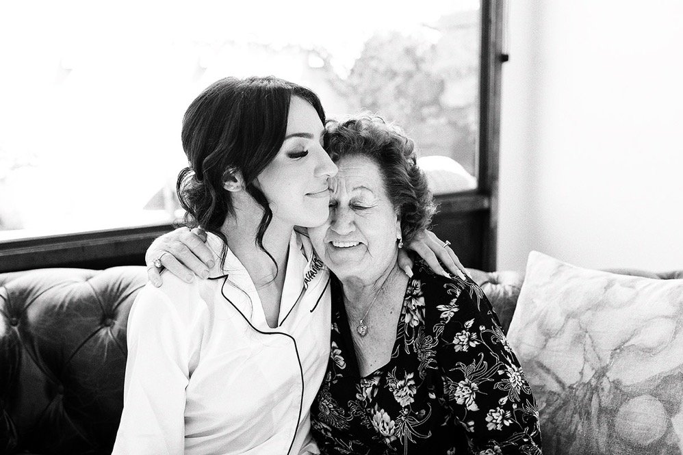 A young woman in a white uniform embracing an elderly woman with curly hair, both with eyes closed and smiling, sitting on a couch near a window.