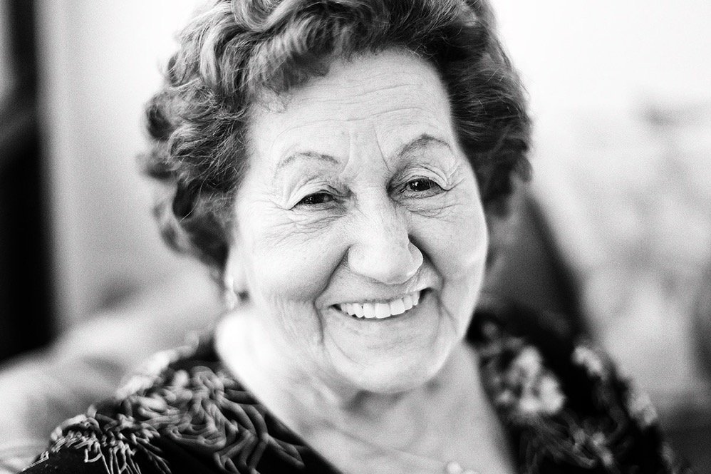 Black and white photo of an elderly woman with short, curly hair, smiling warmly at the camera, wearing a patterned top.