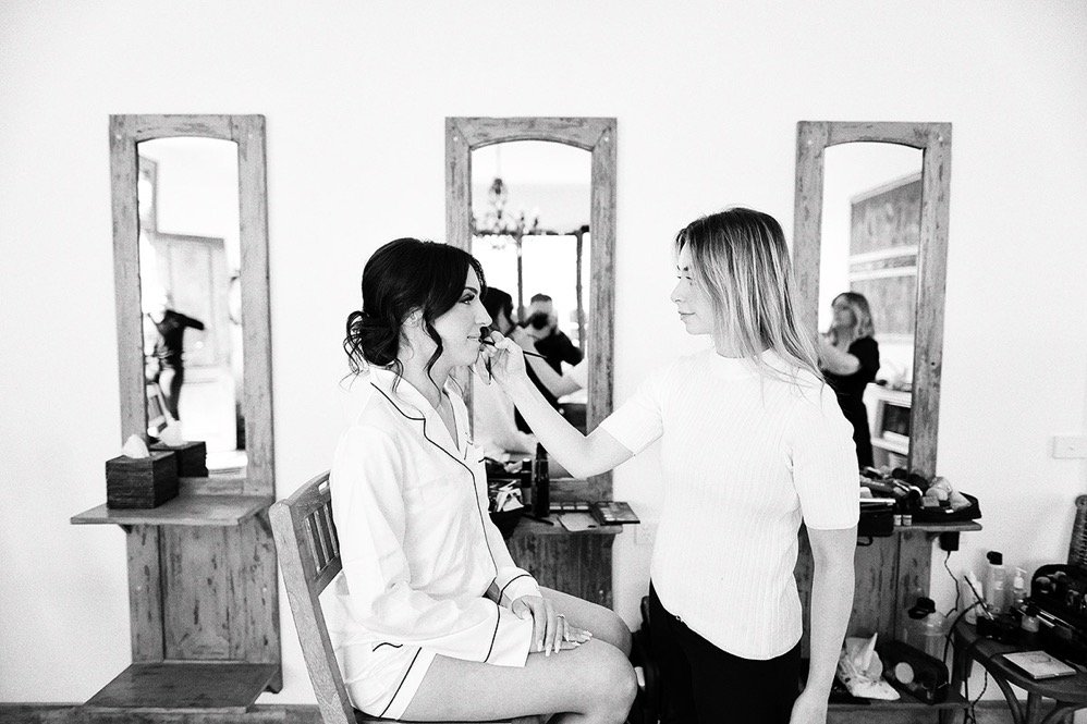 A woman sitting in a salon chair getting her makeup done by another woman in front of three mirrors.