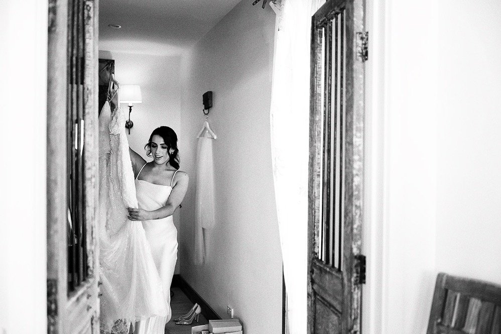 A woman in a white dress holding a wedding gown and smiling in a room with wooden doors, a wall-mounted lamp, and a mirror.