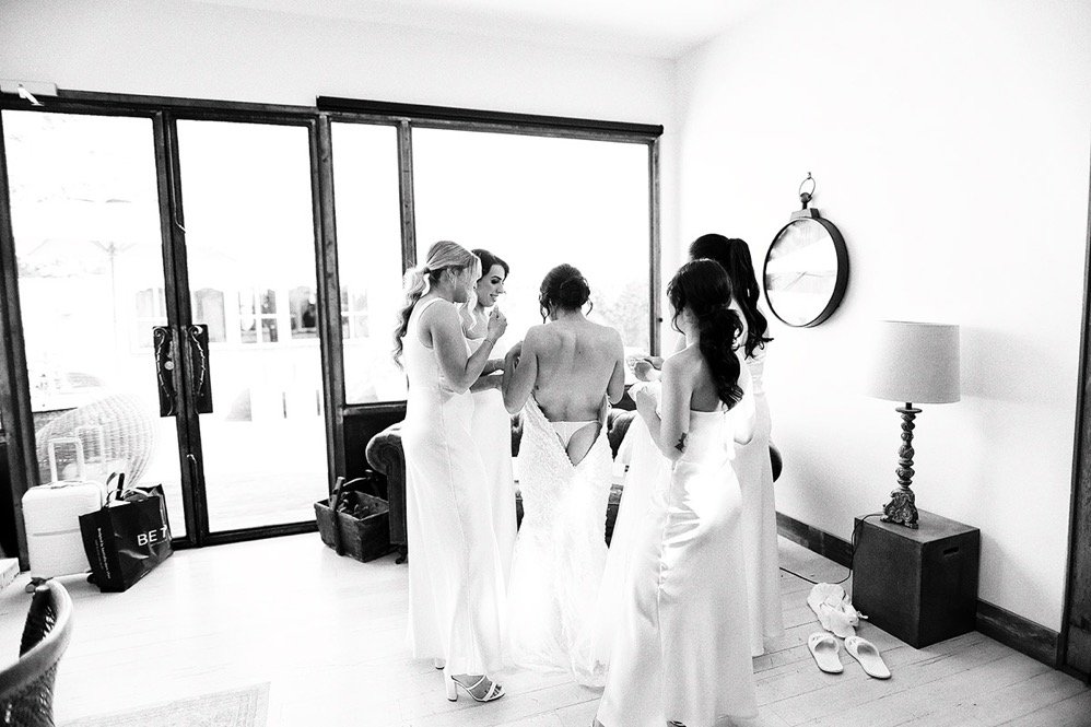 Group of women, some in wedding dresses, gathered in a room with large windows and sliding glass doors, preparing for a wedding.