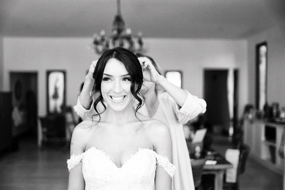 A bride smiling as her hair is being styled in a dressing room, wearing a strapless wedding gown.