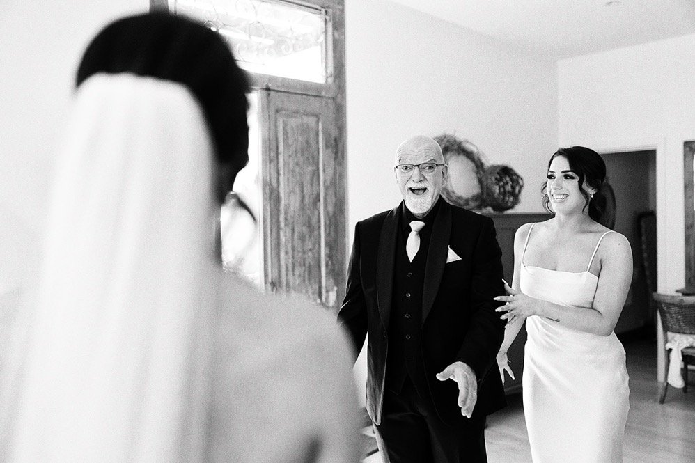 A bride with a veil and wedding dress is looking at an elderly man and a young woman, who are smiling and appear to be in a joyful conversation indoors.