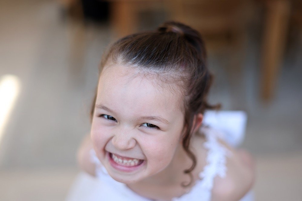 Close-up of smiling young girl with brown hair tied in a ponytail, wearing a white dress, looking up at the camera.