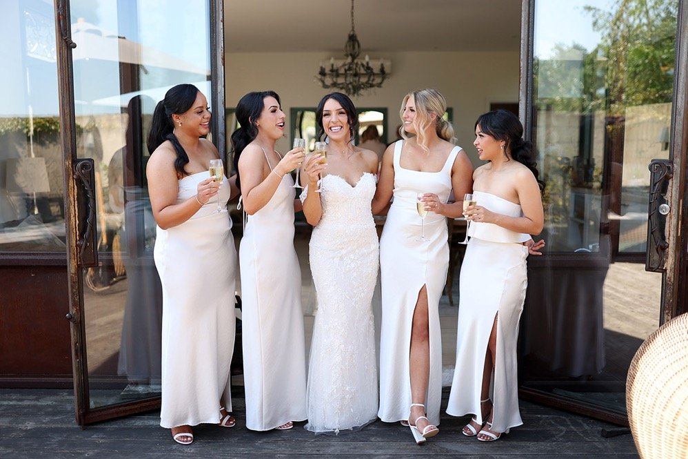 Bride and five bridesmaids celebrating, all in white dresses, holding glasses of champagne, standing near open glass doors in a bright indoor setting.