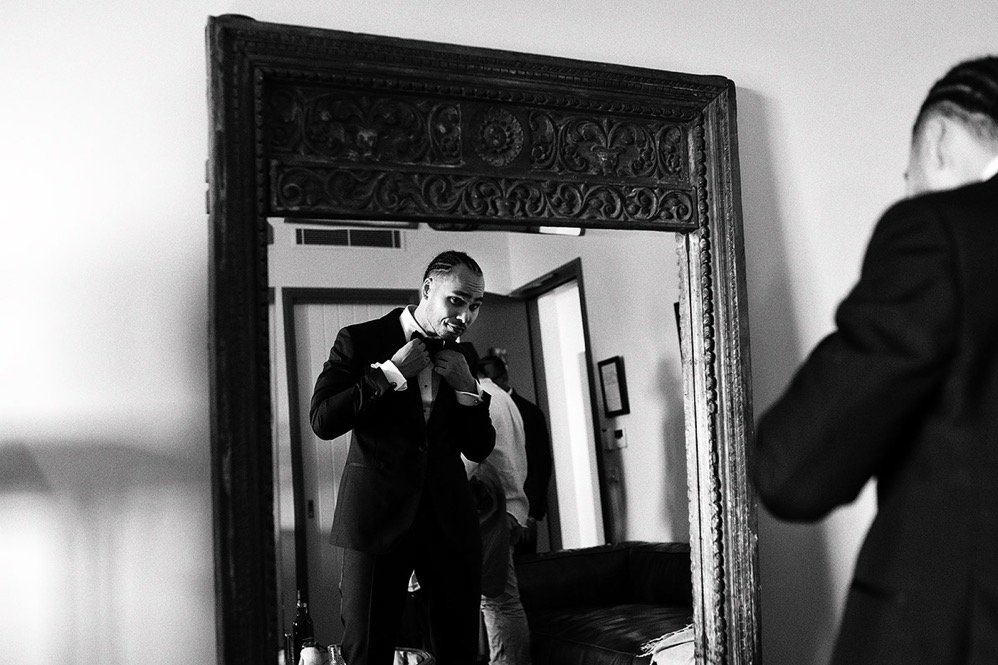A man in a tuxedo adjusting his jacket while looking in a large decorative mirror. The reflection shows another person behind him. The scene appears to be in a room with a door and framed picture on the wall.