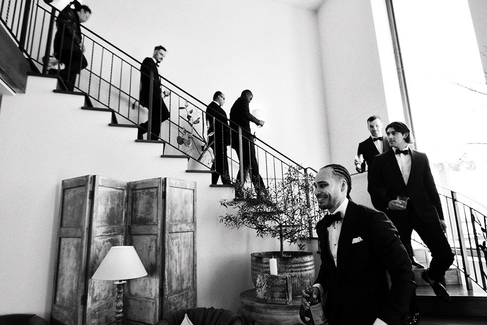 Men dressed in tuxedos walking down a staircase at a formal event, with some holding drinks, in a well-lit modern interior.