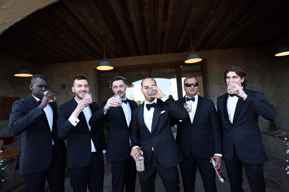 Six men in black tuxedos and bow ties raising glasses for a toast indoors with wooden ceiling and dim lighting.