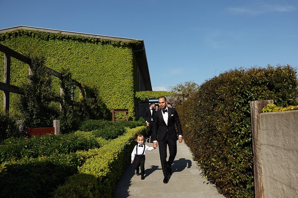 A man in a black tuxedo holding hands with a young boy in a black and white suit, walking on a sidewalk with green hedges and a large green building behind them, under a clear blue sky.