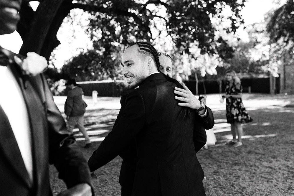 A man with braided hair and a woman are hugging outdoors in a park, smiling. Another person in a floral dress and boy are visible in the background.