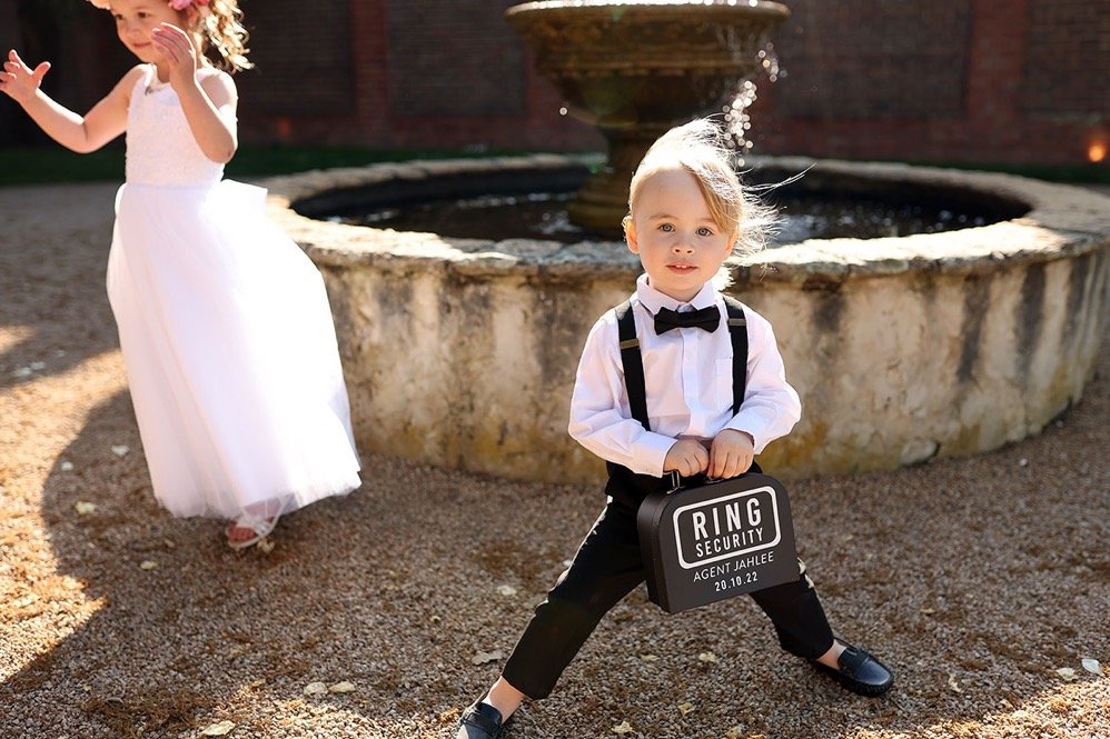 A young boy dressed in a white shirt, black bow tie, and black suspenders holding a suitcase labeled "Ring Security" with nearby children in white dresses near a fountain outdoors.
