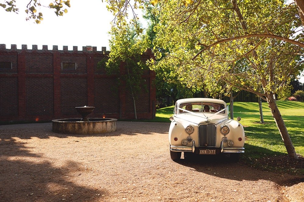 A vintage white car parked under a tree on a gravel area next to a brick wall with a fountain nearby.