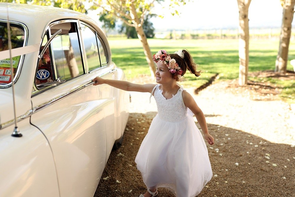 A young girl in a white dress and floral wreath on her head smiling and reaching out to touch a vintage white car parked outdoors on a sunny day, with trees and a grassy background.