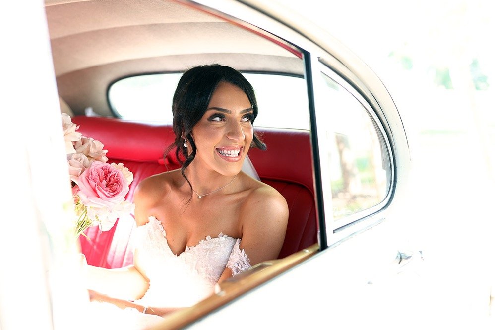 A smiling woman in a wedding dress sitting in the backseat of a car, holding a bouquet of pink roses.