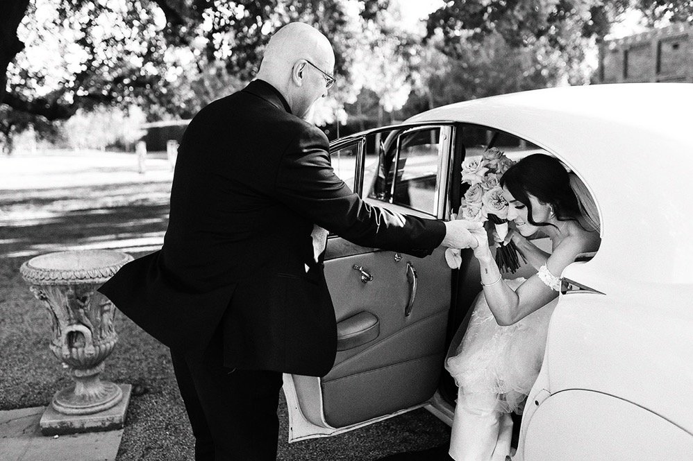 A man dressed in a tuxedo helping a woman in a wedding dress out of a vintage car, with the woman holding a bouquet of roses and smiling.