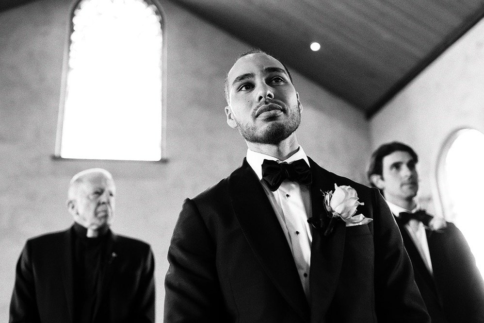 Groom in tuxedo with bow tie and boutonniere during wedding ceremony inside a church, with two men in tuxedos in the background.