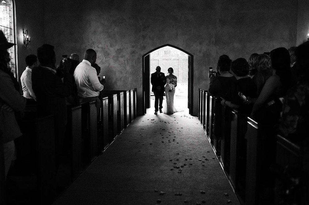 A bride walking down the aisle with a man, likely her father, during a wedding ceremony as guests watch from the pews in a church.