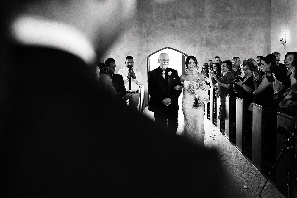 A bride walks down the aisle with her escort at a wedding ceremony inside a church, surrounded by seated guests.
