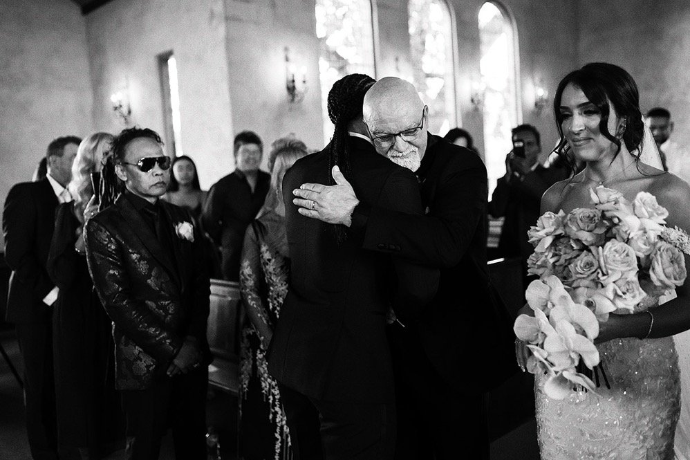 A group of people attending a wedding ceremony in a church, with two men hugging in the center, one of them is an elderly man with glasses. A woman in a wedding dress holding a bouquet of flowers stands to the right, smiling. Others are standing and