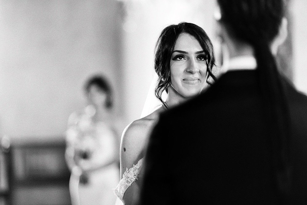 A bride with dark hair and a white dress looks at a person in front of her, possibly during a wedding ceremony. The background is blurred with a person in a white dress standing in the distance.