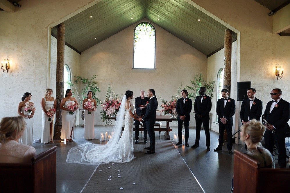A wedding ceremony in a church with the bride and groom holding hands, surrounded by bridesmaids in white dresses holding pink bouquets, and groomsmen in black tuxedos. An officiant is officiating the ceremony, and there are candles and floral arrang