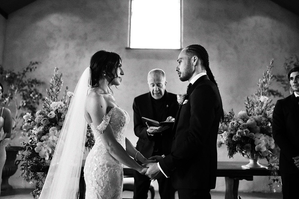 A black and white photograph of a wedding ceremony inside a church. The bride and groom are holding hands and gazing into each other's eyes. The officiant stands between them, reading from a book. Floral arrangements decorate the background.