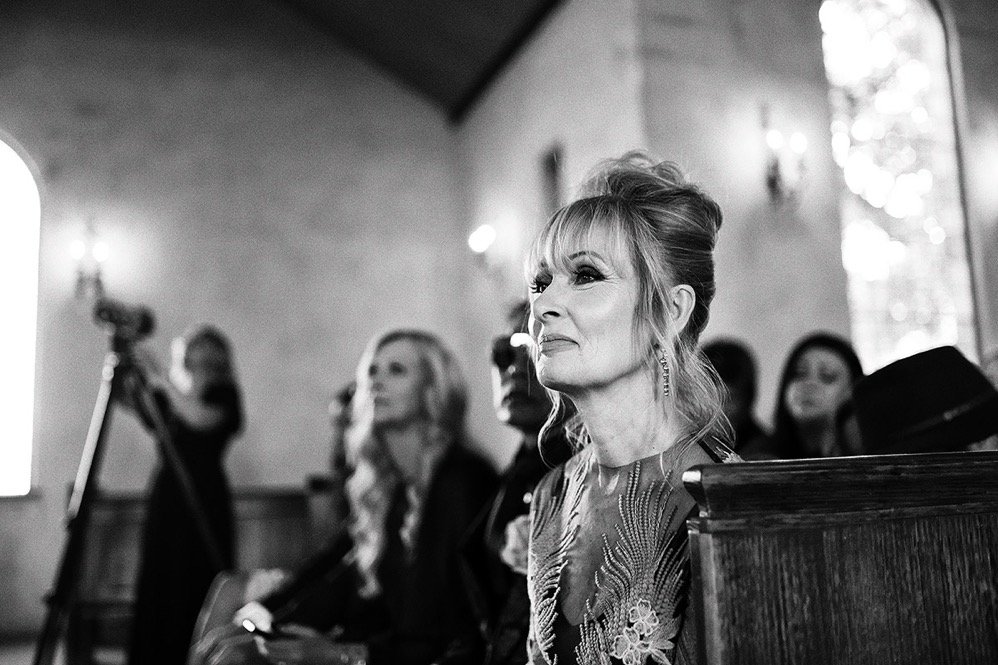 Woman with styled hair and earrings sitting in a church or chapel, seen in black and white, attending a formal event with several other women and a camera on a tripod in the background.