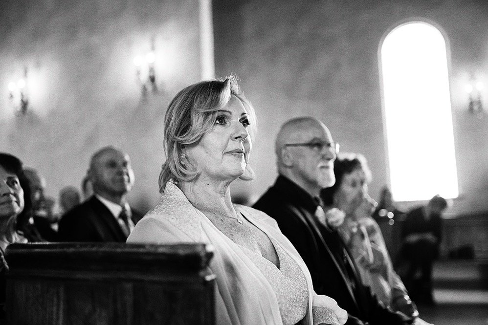 A black-and-white photo of people sitting in a church, focusing on a woman with short, wavy hair in the foreground, with others visible behind her, inside a church with arched windows.