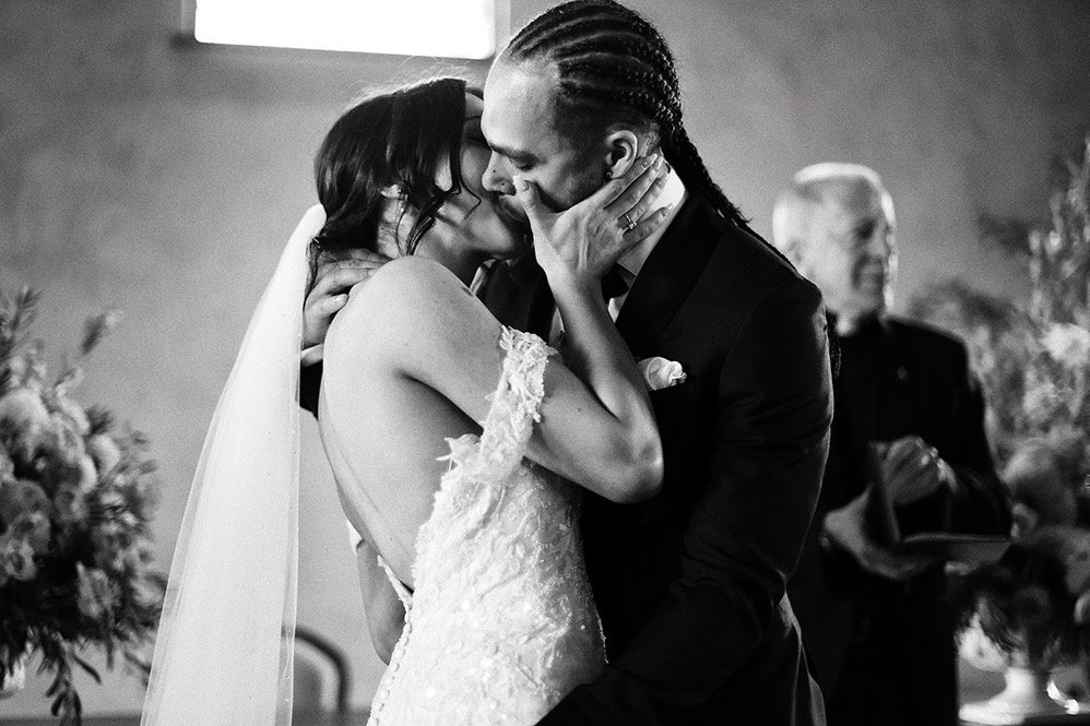 A black and white photo of a bride and groom kissing during their wedding ceremony, with an officiant standing in the background.