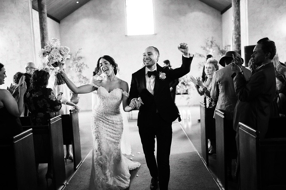 A bride and groom walking down the aisle, smiling and holding hands, in a church with guests clapping and taking photos.
