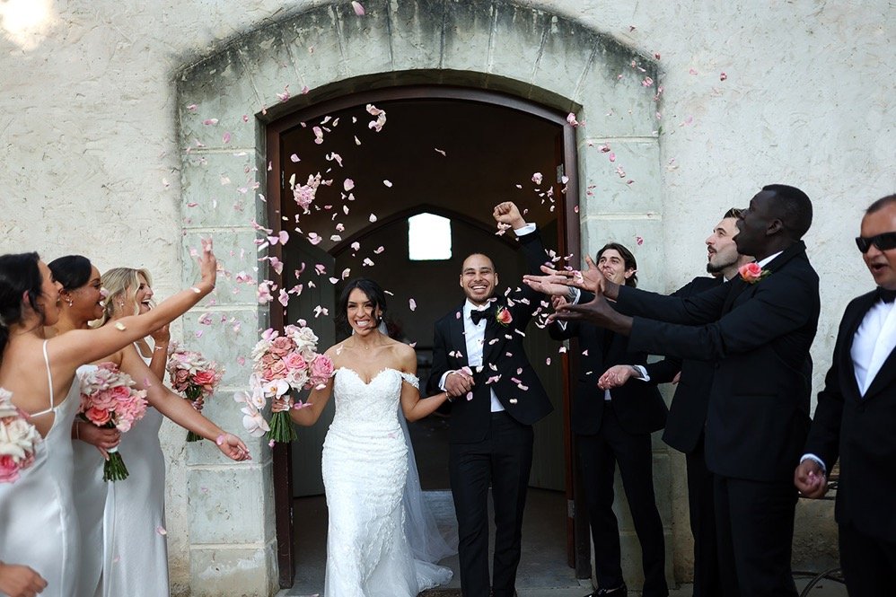 A newly married couple walks out of a building while wedding guests throw pink flower petals in the air.