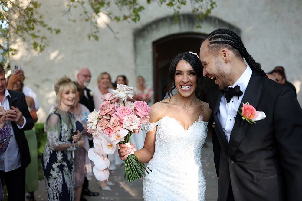 A newlywed couple sharing a joyful moment outside, surrounded by friends and family. The bride holds a bouquet of pink and white flowers, and the groom is dressed in a black tuxedo with a pink boutonniere.