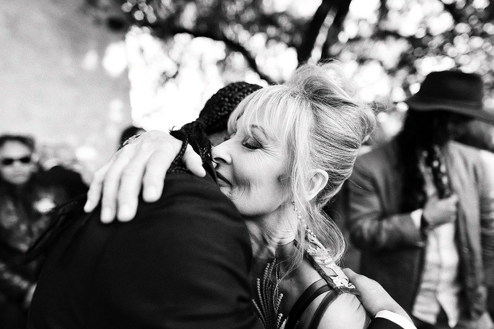 Black and white photo of two people hugging, one woman with light hair and indeterminate ethnicity, smiling with her eyes closed, in an outdoor setting with blurred people and trees in the background.