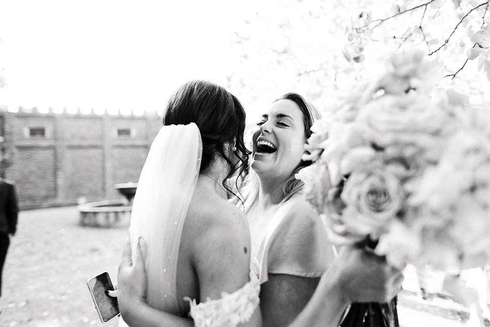 Two women, one with a bridal veil, laughing and embracing outdoors, with a large bouquet of flowers on the right.
