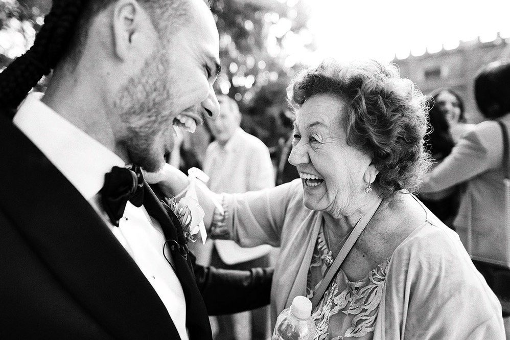 A joyful elderly woman and a smiling man in a tuxedo sharing a happy moment at a social gathering, possibly a wedding, outdoors.
